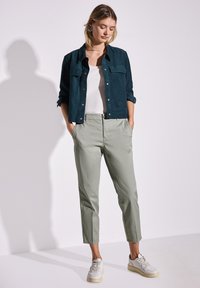 Tmavě zelená semišová zkrácená bunda přes bílý top, spárovaná se světlými zelenými úzkými kalhotami a bílými teniskami s texturovaným detailem.