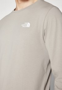 Hellgraues Langarmshirt aus weichem Stoff, mit einem kleinen weißen Logo von The North Face auf der linken Brustseite.