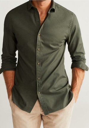 Homme portant une chemise boutonnée vert olive aux manches retroussées et un pantalon en velours côtelé beige, les mains dans les poches, sur un fond clair.