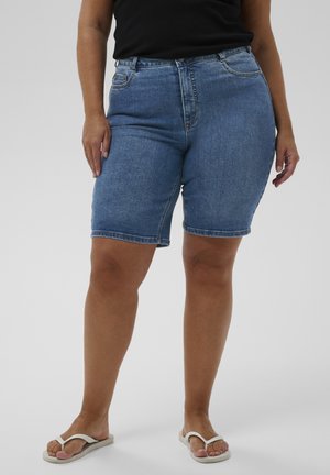 Person iført blå denimshorts til midt på låret, sort top og hvide flip-flops, stående mod en ensartet lysegrå baggrund.