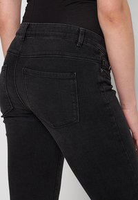 Črne jeans hlače iz denima s prileganjem, opremljene s hrbtnim žepom in gladko teksturo. Pas je strukturiran, primeren za sproščeno nošnjo.