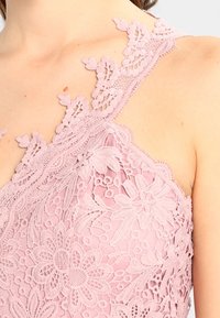 Pehmeän vaaleanpunainen pitsitoppi, jossa kukkakuviot, herkkä aaltoileva reuna ja epäsymmetrinen olkaimien muotoilu.