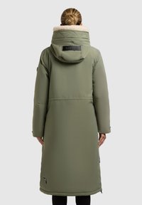 Lang olivengrøn puffermantel med aftagelig hætte, kunstpelsfor, quiltede detaljer og sidelommer. Har mærkning på ryggen.