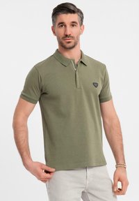 Olivgrön polo t-shirt med korta ärmar, krage och dragkedja fram. Har ett litet svart emblem på bröstet och en slät yta.