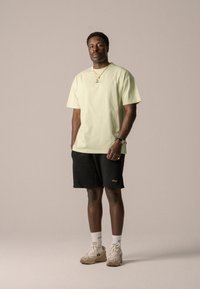 T-shirt en coton vert clair à manches courtes, associé à un short noir. Les accessoires comprennent un collier en or et des baskets beiges avec des accents.