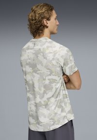 Άνδρας με ξανθά κατσαρά μαλλιά, φοράει κοντομάνικο πουκάμισο με γκρι και λευκό σχέδιο παραλλαγής, στέκεται με τα χέρια σταυρωμένα, κοιτάζοντας πλάγια.