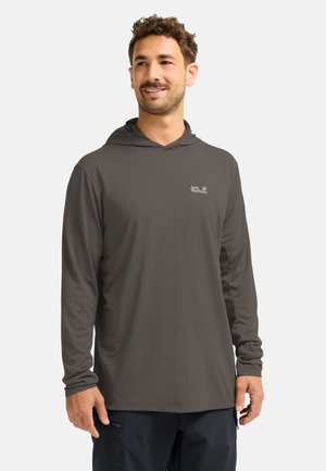 Man draagt een donkergrijze hoodie met lange mouwen en het Jack Wolfskin-logo, staand en glimlachend, tegen een effen lichte achtergrond.