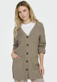 Cardigan en tricot beige avec un col en V, six boutons noirs, deux poches avant et des manches retroussées. Texture douce et coupe décontractée.