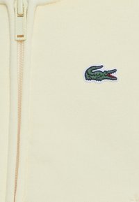 Sudadera amarilla con cremallera, hecha de un tejido suave, que cuenta con un logo bordado de cocodrilo verde en la parte izquierda del pecho. Cremallera parcialmente visible.