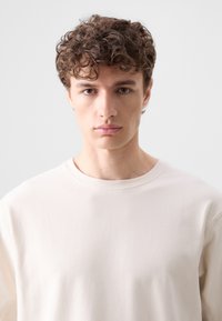 T-shirt z krótkim rękawem, okrągłym dekoltem w jasnym beżowym kolorze z bawełny. Charakteryzuje się gładką fakturą, prostym dołem i luźnym krojem.