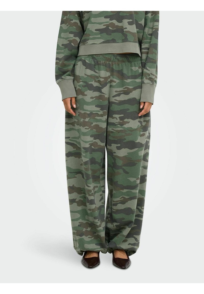 La personne porte un pantalon camouflage ample avec taille élastique et un haut à manches longues assorti, associé à des chaussures pointues foncées.