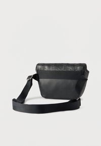 Sac bandoulière noir avec une large sangle réglable, un rabat supérieur texturé et une section inférieure lisse, posé sur un fond clair uni.