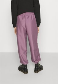 Lila, löst sittande byxor med resårmidja och mudd, tillverkade av ett slätt material. En svart texturerad tröja och svarta stövlar bärs.