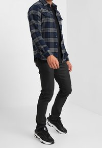 Rutig flanellskjorta i marinblått och grått, med två bröstfickor. Buren över en svart t-shirt, kombinerad med svarta byxor och sneakers.