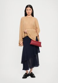 Nainen, jolla on päällä vaaleanbeige kerrostettu paita ja tummansininen epäsymmetrinen hame, pitää punaisesta käsilaukusta kiinni ja hänen jalkoihinsa kuuluu mustat loafersit, joissa on kultaketjuyksityiskohta.