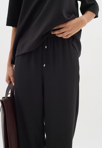 Černé volné kalhoty s úzkým pasem a šňůrkou, zakončené kovovými konci, v kombinaci s oversized černým topem. Je držena tmavá kabelka.