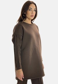 Brauner, übergroßer Pullover mit langen Ärmeln, Rundhalsausschnitt und seitlichen Schlitzen. Glatte Textur mit kontrastierenden vertikalen Nähten an den Ärmeln.