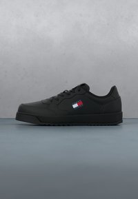 Baskets basses noires avec laçage à l'avant et logo Tommy Jeans en rouge, blanc et bleu sur le côté, sur fond gris.