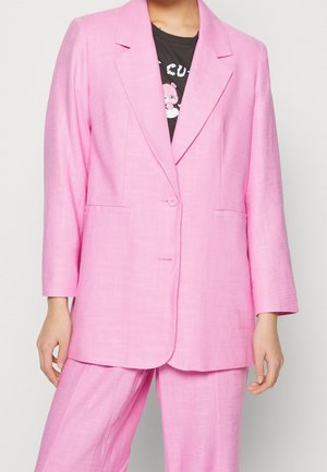 Mujer con blazer fucsia brillante y pantalones a juego, luciendo una camiseta gráfica negra con un diseño de personaje en color rosa.