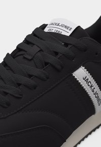 Μαύρο αθλητικό παπούτσι με υφασμένα μαύρα κορδόνια, λευκή ετικέτα στη γλώσσα με την ένδειξη "JACK&JONES EST. 1990" και λευκή πλευρική λωρίδα με το όνομα της μάρκας.