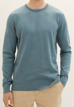 Homme portant un pull bleu à col rond avec poignets et ourlet côtelés, associé à un pantalon beige, une main dans la poche sur un fond uni.