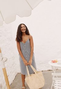 Robe à rayures avec des boutons sur le devant, dotée de bretelles spaghetti. Le modèle tient un sac en paille tressée. L'arrière-plan est un mur blanc texturé.