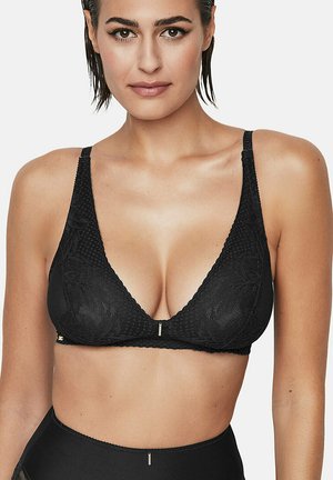 Vrouw in een zwarte kant bralette met dunne bandjes en een bijpassende high-waisted onderkant tegen een eenvoudige witte achtergrond.