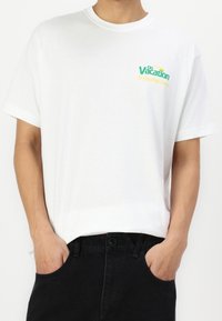 Bílá bavlněná tričko s krátkými rukávy, s nápisem "Na dovolené" v zelené a žluté barvě na hrudi, kombinováno s černými denimovými kalhotami.