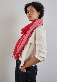 Rød og lyserød frynset tørklæde draperet over en lysblomstret bluse, der har en blød tekstur og et lagdelt design, mod en neutral baggrund.