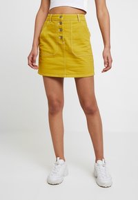 Mini-jupe en denim jaune à taille haute, avec un devant à boutons et deux poches avant. Associée à un crop top blanc uni et des baskets blanches.
