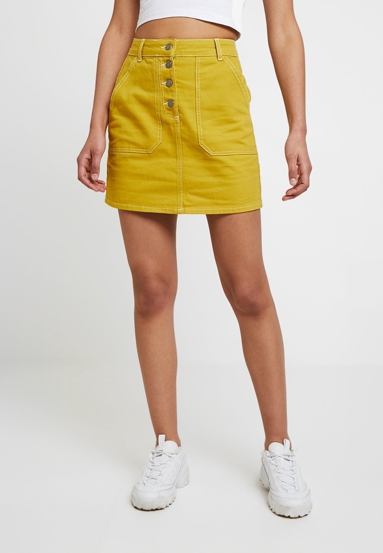 Mini-jupe en denim jaune à taille haute, avec un devant à boutons et deux poches avant. Associée à un crop top blanc uni et des baskets blanches.