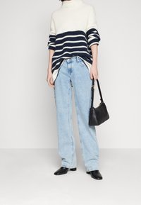 Mujer con un suéter a rayas blancas y marinas, jeans de tiro alto en azul claro, botas negras hasta el tobillo, sosteniendo un pequeño bolso negro.