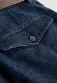 Tissu texturé bleu foncé avec une poche à rabat boutonnée et des coutures visibles, faisant partie d'un vêtement avec une ceinture marron en haut.