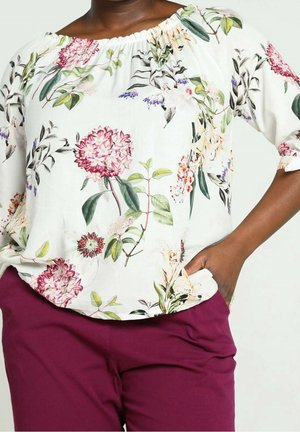 Kvinde iført en hvid bluse med bare skuldre med pink og grønne blomsterprint og lilla bukser, med hånden i lommen, stående mod en ensfarvet baggrund.