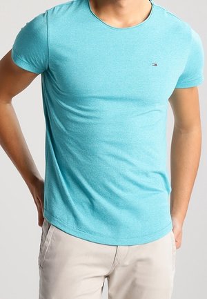 Młody mężczyzna w dopasowanej turkusowej koszulce i jasnych spodniach, stojący z rękami na biodrach na neutralnym tle.