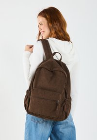 Sac à dos en velours côtelé marron avec un haut arrondi, une poche zippée à l'avant et des bretelles réglables, doté de poches en maille sur les côtés pour un espace de rangement supplémentaire.