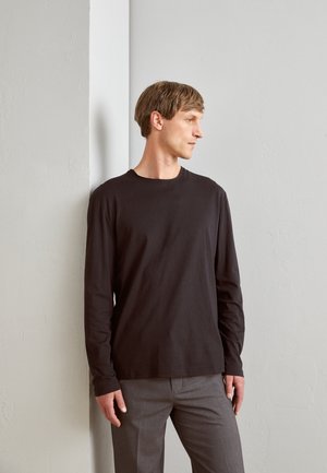 Schwarzes Langarm-T-Shirt aus weichem Stoff, mit Rundhalsausschnitt und lockerer Passform, kombiniert mit grauen Hosen, vor einer hellen Wand.