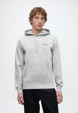 Jeune homme aux cheveux bouclés portant un sweat à capuche gris clair avec un petit logo noir « Lacoste » sur la poitrine et un emblème de crocodile sur la manche.