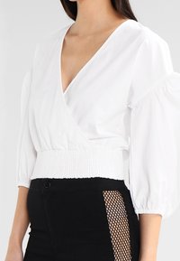 Vit omslagstop med djup V-hals och voluminösa puffärmar, med en smockad midja. Kombinerat med en svart kjol som har ett meshnät-panel.