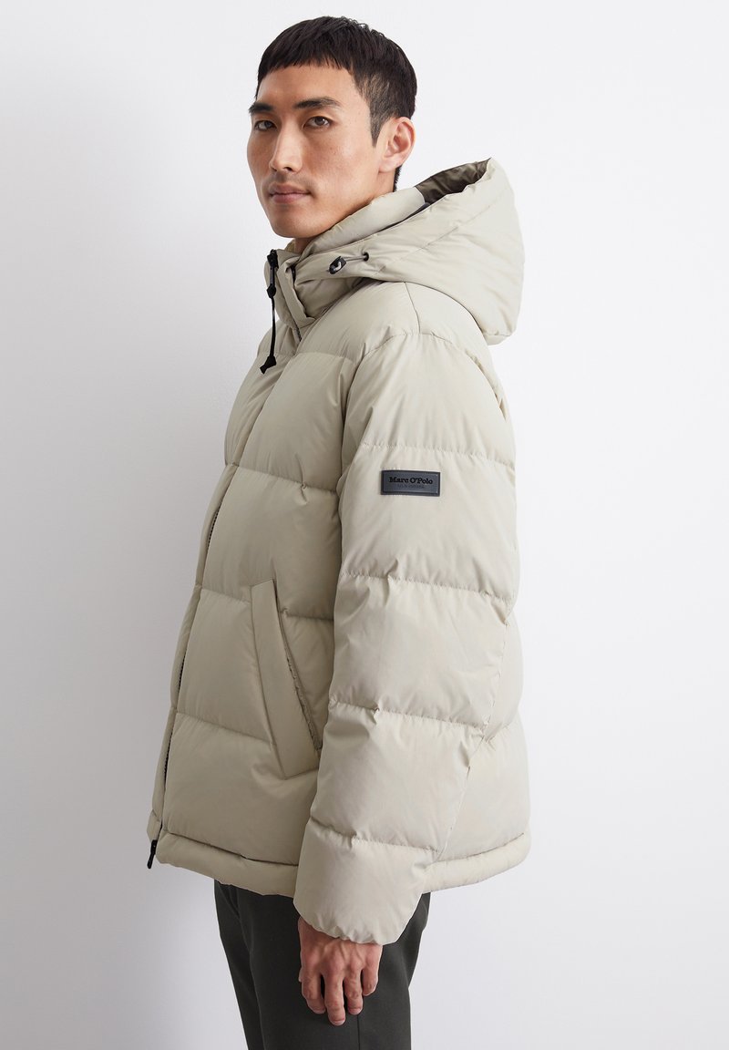 Beige Daunenjacke mit Kapuze, gestepptem Design und schwarzen Akzenten. Sie verfügt über seitliche Taschen und ein Logoschild auf dem linken Ärmel.