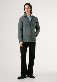 Un joven se presenta con confianza en una chaqueta de cuadros y pantalones negros, con un fondo neutro que acentúa su aspecto casual pero con estilo.