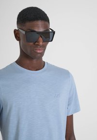 Gafas de sol rectangulares negras con lentes oscuros, llevadas con una camiseta de manga corta azul claro que presenta un pequeño logo en el lado izquierdo del pecho.