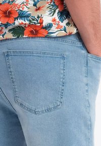 Un jean en denim bleu clair présente une grande poche arrière avec un bord cousu. Le tissu a une texture lisse et un léger dégradé.