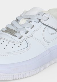 Primer plano de una zapatilla blanca con puntera perforada, cordones gruesos, logo Nike swoosh y una tira de velcro sobre los cordones.