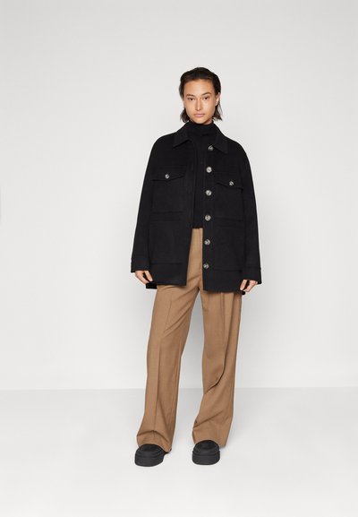 Schwarze Außjacke mit Kragen, Fronttaschen und Knopfschluss, kombiniert mit beigefarbenen, weit geschnittenen Hosen und dunklem Schuhwerk.
