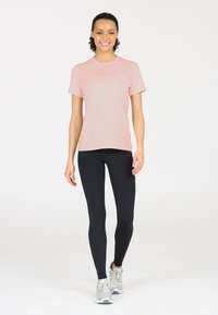 Vaaleanpunainen lyhythihainen urheilupaita, jossa on sileä tekstiili, yhdistettynä tiukkoihin mustiin legginsseihin ja vaaleanharmaisiin lenkkareihin neutraalilla taustalla.