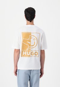 Hvid bomulds t-shirt med et stort gult grafisk design på ryggen, med "HUGO" tekst og geometriske former. Korte ærmer, rund hals.