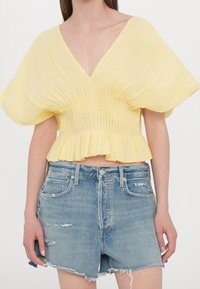 Helle gelbe kurze Bluse mit tiefem V-Ausschnitt, plissierter Textur, weiten kurzen Ärmeln und einem fransigen unteren Saum, kombiniert mit zerrissenen Jeansshorts.