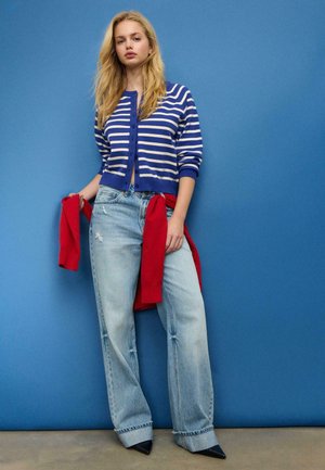 Oprijeta modro-bela črtasta jopica, ohlapne svetlo modre obrabljene kavbojke, rdeč pulover, ki je povezan okoli pasu, in ostri črni čevlji z visoko peto na modri podlagi.