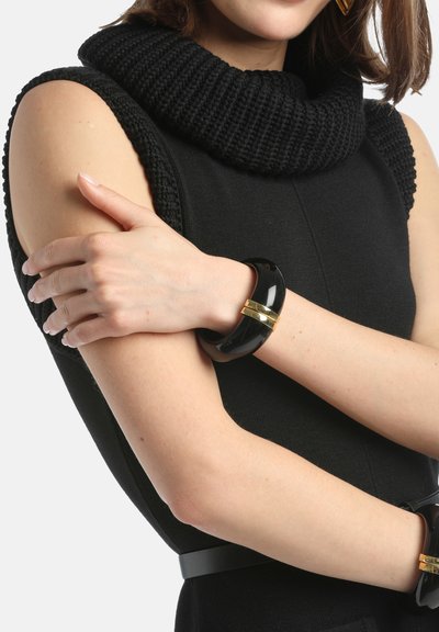 Czarny sweter z dekoltem typu cowl, dopasowany w talii, z błyszczącą czarną plastikową bransoletką ze złotymi akcentami na nadgarstku.